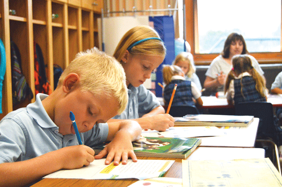 In the first- and second-grade classroom at Mary, Queen of Peace School in Rogers, Joe Macke and Addison Hynes work on an assignment in the foreground while teacher Jennifer Bolduc works with other pupils in the background.