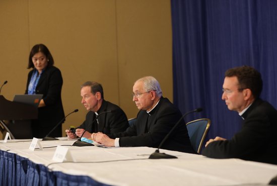 Bishop Michael F. Burbidge of Arlington, Va., Archbishop José H. Gomez of Los Angeles, president of the U.S. Conference of Catholic Bishops, and Auxiliary Bishop Andrew H. Cozzens of St. Paul and Minneapolis, attend a Nov. 16, 2021, news conference during the bishops' fall general assembly in Baltimore. At the podium is Chieko Noguchi, USCCB director of public affairs.