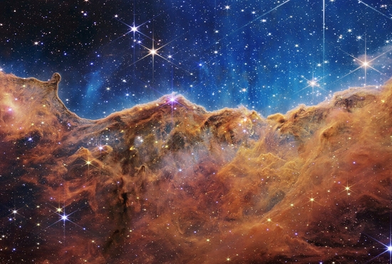 The "Cosmic Cliffs" of the Carina Nebula are seen in an image released by NASA released July 12, 2022. The "cliffs" are divided horizontally by an undulating line between a cloudscape forming a nebula along the bottom portion and a comparatively clear upper portion. The image is from data provided by NASA's James Webb Space Telescope, a revolutionary apparatus designed to peer through the cosmos to the dawn of the universe. Speckled across both portions is a starfield, showing innumerable stars of many sizes.