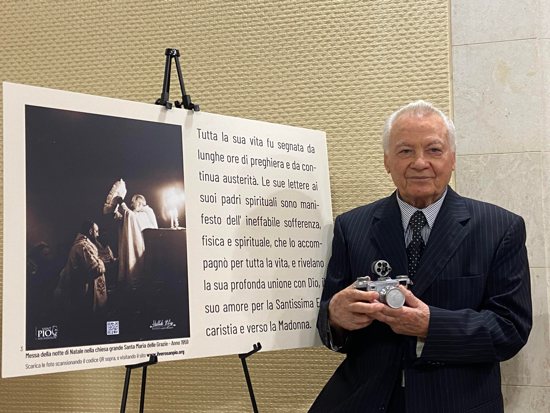Elia Stelluto, St. Padre Pio's personal photographer, stands before a poster showing a previously unseen photo of the saint while holding the camera he used to capture it during a presentation at the Vatican April 29, 2024.