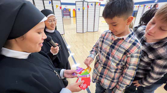 Pro Ecclesia Sancta Sister Fabiola Valdeiglesias hands out Holy Cards to Joaquin Canchaya while Oliver Linder looks on and PES Sister Emy Ychikawa holds a microphone during Catholic Schools week in January 2023 at St. John the Baptist in Savage.