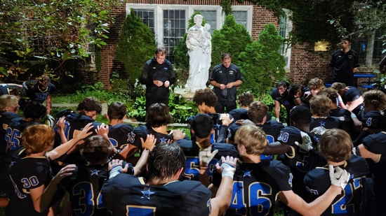 At left, Father Michael Tix gathers with the Holy Angels varsity football team in the school courtyard — near a statue of St. Joseph, commemorating the Sisters of St. Joseph who founded the Richfield school — for prayer and comments by the head coach.