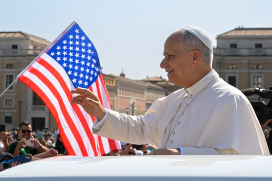 With a U.S. flag in the background, Pope Leo XIV waves to the crowd from the popemobile as he rides around St. Peter's Square at the Vatican before his weekly general audience Aug. 6, 2025.